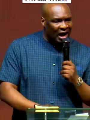 A man wearing a blue checked collared shirt speaks into a black microphone, his mouth open, with a red background. The video is brightly lit.