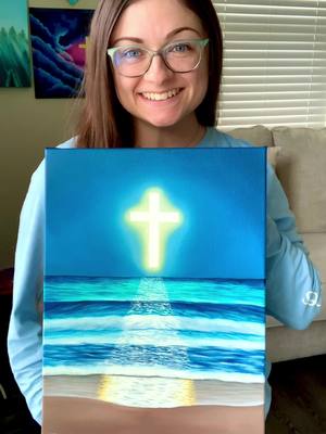A woman with glasses smiling, holding a painting of the ocean with a glowing cross in the sky, a sign of faith.