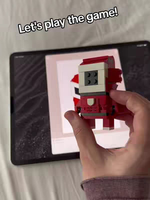 A hand holding a red and white Lego figure against a black tablet displaying a matching image. The background is a white bed sheet.