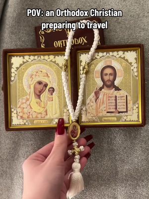 A hand holding prayer beads and small icons of religious figures, set against a background that includes an open book and a plain, grey surface.