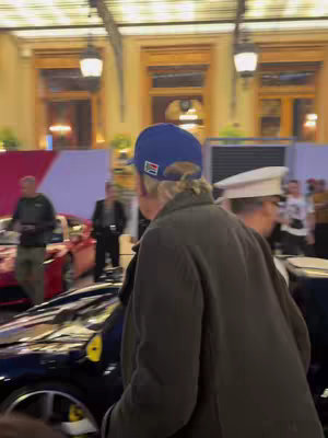 An individual with his back to the camera stands near a dark blue Ferrari. He is wearing a blue cap and an olive-green coat. In the background, people are present, and the setting appears to be at a public event with high-end cars.