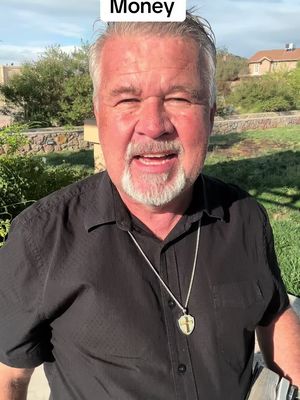 The cover image features a middle-aged man with a grey beard and short hair, smiling directly at the camera. He wears a black shirt and a necklace with a pendant. Green foliage and a blurred outdoor setting form the background, with a partial view of a building.