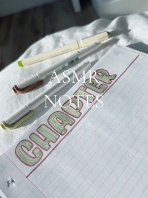 A close-up shot of a notebook page with the word "CHAPTER" written in large, green bubble letters with a red outline. Three pens are placed above the notebook. The background is a white surface, possibly a bedsheet, and natural light is illuminating the scene.