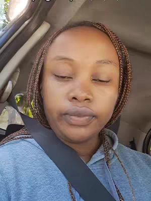 A close-up shot of a woman inside a car. She has medium-brown skin, wears brown braids, and a gray hoodie, with a seatbelt across her shoulder. Her eyes are closed, suggesting reflection or a moment of thought. The car's interior, including the seat and door, is visible in the background, which indicates the setting.
