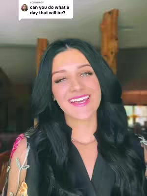 A woman with long black hair is singing with a smile. She is wearing a black outfit with floral sleeves, likely in a living room setting. The background includes wood trim and a glimpse of a window.