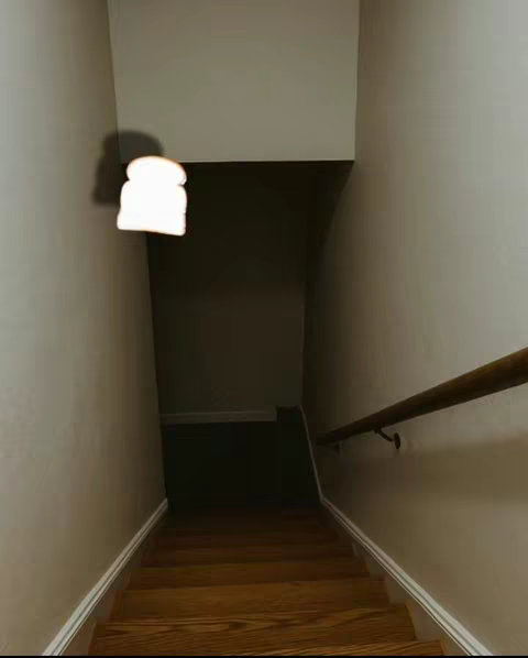 A slightly low-angle shot down a flight of wooden stairs. The walls are a light beige, and a wooden handrail runs along the right side. A slice of white bread is hovering in the air near the top of the stairs casting a shadow, as if it is floating down the staircase.