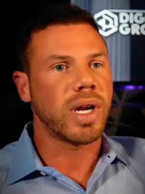 A man with light skin is the central focus, his face filling most of the frame. He has short, dark hair, blue eyes, and a neatly trimmed beard and mustache. The man is looking at the viewer with a slightly open mouth as if speaking. He is wearing a light blue collared shirt. The background is dark, with a blurry logo visible to the upper right.