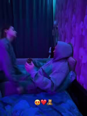 A dimly lit room features two figures. The focus is on a person wearing a gray hoodie, seated in a bed. The background includes blue and purple lighting and part of a bed structure.