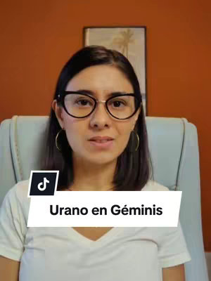 A woman with glasses is looking directly at the camera, with an orange-colored wall as the background. A black and white banner that says "Urano en Géminis" sits at the bottom.
