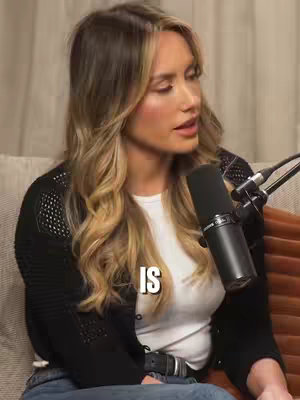 The cover image shows a woman seated, turned slightly to the side. She has long, wavy blonde hair. She is wearing a white top under a black, open-knit cardigan and blue jeans. A microphone is positioned near her mouth. The word "IS" in a stylized white font is overlaid on her chest. In the background, there is a textured beige wall.