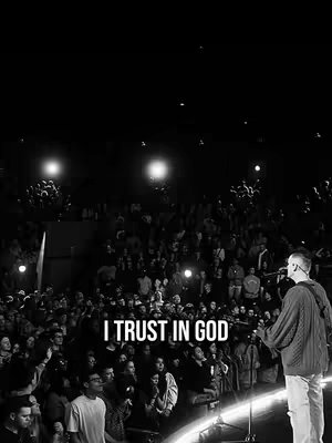 A black and white image of a live performance. A crowd is visible, with a person singing into a microphone on a stage lit by spotlights. The text "I TRUST IN GOD" is overlaid on the image.