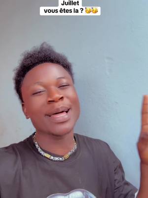A young man with dark skin is centered in a medium shot. He has short, styled black hair and is wearing a black t-shirt and a silver necklace with gold accents. His facial expression is animated, and he is gesturing with one hand raised, as if making a point. Above him, text in white reads 'Juillet vous êtes la ?' 