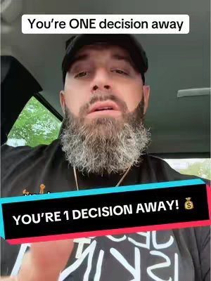 A man with a beard and a black cap is sitting inside a car, looking directly at the camera. Text overlays appear above and below him. The upper text reads, "You're ONE decision away," while the lower text, on a colored bar, reiterates, "YOU'RE 1 DECISION AWAY!" and includes a money bag icon. The man's expression shows a slight focus, and the background, including the car's interior and some greenery, is slightly blurred.