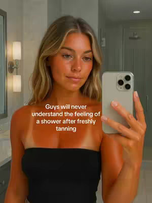 A young woman stands in front of a mirror, taking a selfie with her phone. She has long blonde hair and is wearing a black strapless top. Her skin has a noticeable tan with clear tan lines on her chest. The background shows a well-lit bathroom with a mirror and sconces.