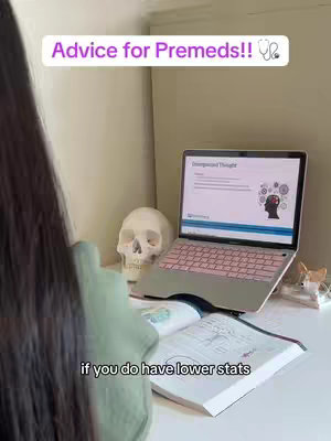 The image shows a desk setup. A woman is seen from the back, her hair covering her face. On the desk is an open book with diagrams, a laptop displaying a presentation, and a skull. The laptop is positioned towards the right with a pink keyboard and screen displaying a presentation. The background is a soft, neutral color.