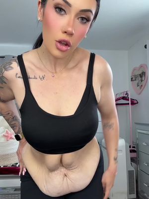 A woman wearing a black sports bra and black pants is shown from the waist up. Her abdominal area is prominent, featuring loose skin. She has makeup on, including blush, and her dark hair is pulled back. The background includes blurred elements suggestive of an interior setting.