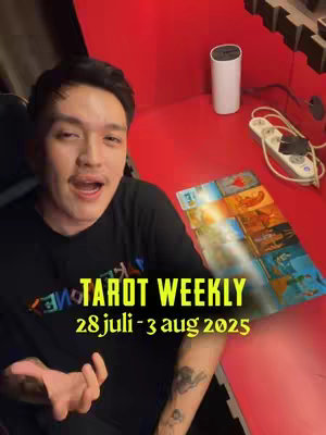 A man with short dark hair wearing a black shirt is in a room with a red background. The man is speaking with his mouth open wide. In the foreground are tarot cards. A text overlay on the bottom of the image says "TAROT WEEKLY 28 juli-3 aug 2025".