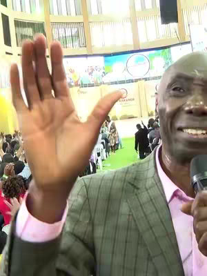 A middle-aged man with a bald pate is smiling, raising his right hand towards the camera. He's wearing a gray and green plaid blazer over a pink collared shirt. Various people and architectural elements form the backdrop. A microphone is held close to his face.