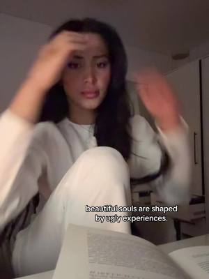 A woman in a white outfit is seated, touching her hair with both hands and glancing downward. The image has a soft, natural aesthetic. Below, a book is open. A text overlay reads, "beautiful souls are shaped by ugly experiences."