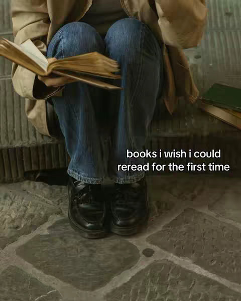The cover image shows a person sitting on a stone surface with their legs crossed and a book in their hands. The person is wearing jeans, black shoes, and a tan jacket. There is a stack of books on the right. The text overlay says: books i wish i could reread for the first time.