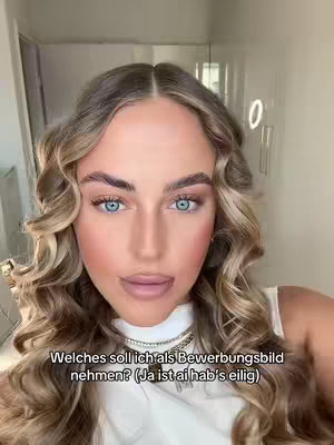 A close-up shot of a young woman with long, wavy blonde hair and light blue eyes, looking directly at the camera. She has full makeup on, including defined eyebrows, blush, and a nude lipstick. A shiny silver necklace is visible. The background is blurred, suggesting an indoor setting with a door and a lit circular object.