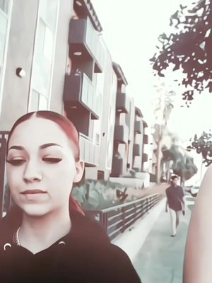 A young woman with red hair is framed from the chest up, looking downwards with a neutral expression. She is wearing a black hoodie and a gold necklace. The background includes a paved walkway and a tall building on the right, with someone walking towards the left.