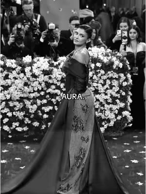 A black and white photo capturing Kylie Jenner at an event, likely the Met Gala. She is the central focus, posing in a flowing, elegant gown with a dramatic train. The background is slightly blurred, featuring photographers and other attendees. The overall composition emphasizes Jenner's poise and the high-fashion environment.
