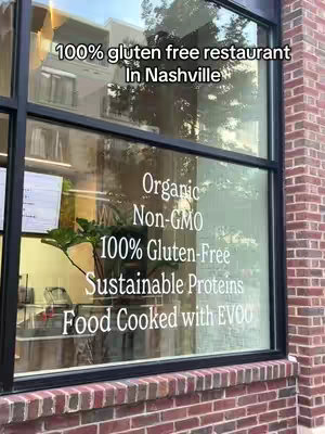 Exterior shot of a restaurant window, with signs visible about organic, gluten-free food, and sustainable practices. The window reflects trees and the building across the street.