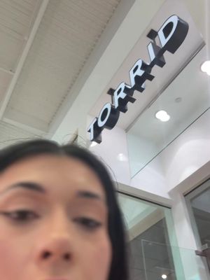 A selfie-style image with the creator's face in the foreground, slightly out of focus. In the background, the storefront of a TORRID store is visible, with the store's sign prominently displayed. The lighting appears to be from an interior mall setting, with a neutral ceiling and recessed lighting.
