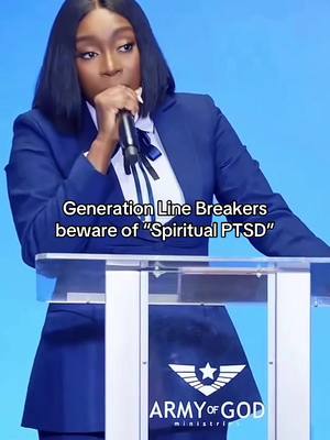 An individual, presumed to be a woman, is speaking at a podium. A microphone is held close to her mouth, with a direct gaze suggesting she is addressing an audience. The background is a plain, light blue. She is wearing a navy blue suit with a white undershirt and a black tie.