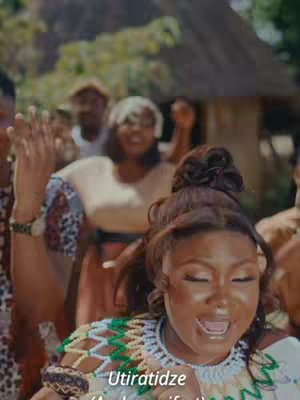 A woman with dark skin and a styled updo is the focal point, looking up and to the right with an open mouth as if singing or speaking. Additional people are visible in the background, with a crowd-like feel.