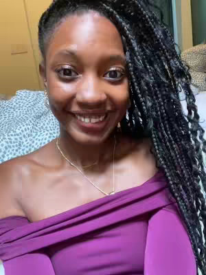 A selfie-style shot featuring a Black woman with long box braids. She's wearing a plum-colored off-the-shoulder top and a gold chain necklace. The background shows glimpses of a bed with a patterned comforter. She smiles, displaying her teeth, and has a warm, direct gaze.