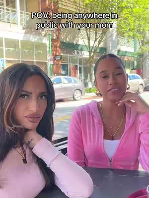 Two women are seated outdoors at a table, with a blurred background of a street scene. The woman on the left is dark-haired with a serious expression, resting her chin on her hand. The woman on the right has lighter hair, is wearing a pink zip-up jacket and has a slightly open smile. Overhead text gives context.