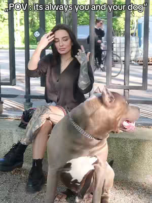 A woman with long dark hair sits on a concrete step with a large dog. The background includes green trees, a metal fence, and a person riding a bicycle. The woman is wearing a dark shirt and jeans, and the dog has a thick, light-colored coat.