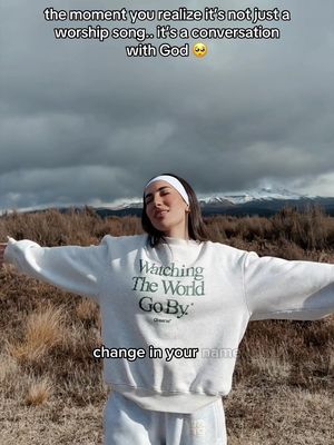 The image shows a woman standing outside with her arms outstretched, a serene background of mountains and a cloudy sky behind her. She is wearing a white sweatshirt, a white headband and has her eyes closed. The top includes text that says "the moment you realize it's not just a worship song... it's a conversation with God". Below that it says "change in your name".