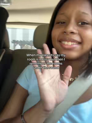 A selfie taken from the front seat of a car. A young Black woman with dark hair and a light blue shirt smiles broadly, waving her hand towards the camera. The background shows the interior of the car and an out-of-focus view of the outside world. White text is overlaid at the bottom of the visible part of the screen.