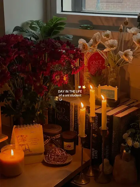 The image showcases a cozy, warm indoor scene with a variety of objects on a table or desk. A window is visible in the background, letting in diffused natural light. Several candles in different sizes are lit, casting a warm glow and creating soft shadows. There are books, some with visible titles, arranged in a stack, indicating a focus on academia or reading. A small calendar, flowers, and decorative items like jars and a decorative metallic sphere add to the aesthetic and theme of a university student's life.