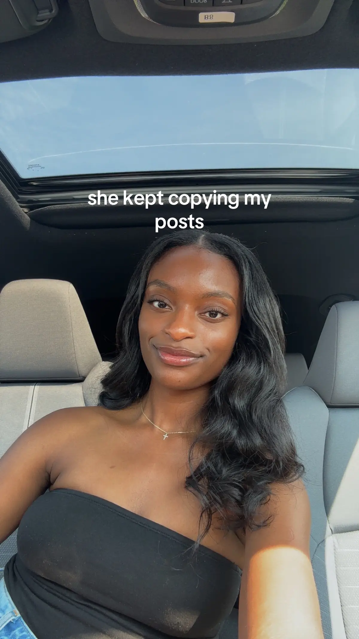 A selfie taken from inside a car, with a young woman in the front seat. The woman is wearing a black strapless top and a necklace with a small cross pendant. Daylight streams in through the sunroof, illuminating her face and hair, which is long and wavy. She is smiling slightly, and the car's interior is visible in the background.