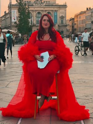 A woman with red hair and a red dress is sitting on a chair in a public square. She is wearing a long, flowing red shawl and holding a piece of paper. The background shows a building with architectural details and other people in the square. The lighting is natural, suggesting an outdoor setting.