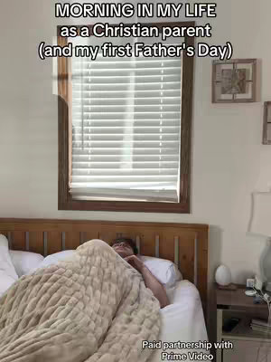 A bedroom scene as seen in a low angle, with the person in bed under a blanket which covers them mostly.  A large window with blinds is centered in the background, framed by brown wooden trim. A small square framed picture and a bedside table are also visible. The lighting appears to be warm and natural.