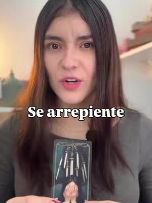 A woman with long dark hair is centered in the frame, looking directly at the camera. She's speaking, with a concerned or questioning expression. In her hands is a tarot card, featuring imagery of swords and a hooded figure. Behind her is a blurry background. The image is well-lit.