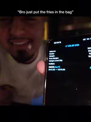 A man is holding a smartphone close to the camera, displaying a trading app interface. The man is smiling and part of his face is in the frame; behind him is a blurred background.