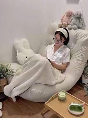 A cozy shot of a person sitting on a large, curved, cream-colored beanbag chair, holding a phone. They are covered with a light-colored blanket, and a plush bunny is on the left side of the chair. A small wooden tray with a drink and a plate with a green item sit in front of the person. In the background are a plant, some stuffed animals, and a wall.
