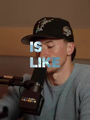 A man wearing a black cap and a gray sweater, with the text 'IS LIKE' overlaid on his face. A microphone is in front of his face.