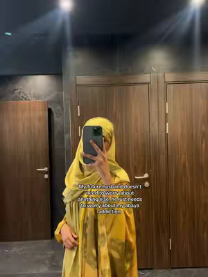 A woman in a yellow abaya and hijab takes a mirror selfie in a public restroom. She is holding her phone up to capture the image. The background features brown wooden doors and a dark ceiling. Text is overlaid on the image in a white font.
