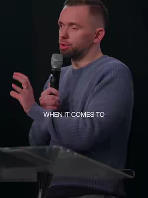 A man with short brown hair, wearing a blue sweater, stands behind a podium and speaks into a microphone. The image is set on a dark background. The text "WHEN IT COMES TO" is at the bottom of the image.