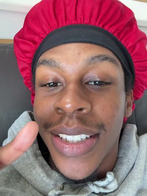 A selfie-style shot featuring a dark-skinned man with a red durag. His face is centered, with a close-up view of his eyes, mustache, and a wide smile that reveals white teeth. He gives a thumbs-up with his left hand. The background is minimalistic, with a glimpse of a neutral-toned wall.