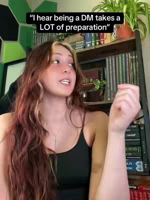 A woman with long, wavy auburn hair is speaking. She's wearing a black tank top and a silver cross necklace. The background includes a bookshelf filled with books. A text overlay reads, "I hear being a DM takes a LOT of preparation."