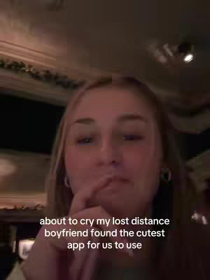 A medium shot features a young woman with long blond hair, gazing towards the camera with a slightly concerned expression. She has light skin. Overlayed below the woman is text stating the app, which she is using for a long distance relathionship. The background, slightly blurred. There's a hint of a decorative element, suggesting an indoor setting with warm lighting.
