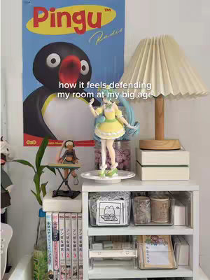 A well-lit room corner shows a shelf with various items: manga, a lamp, figurines, and decorative storage. A poster with a cartoon penguin character labeled 'Pingu' is visible on the wall. The color palette is soft with whites and pastels, creating a cozy aesthetic.